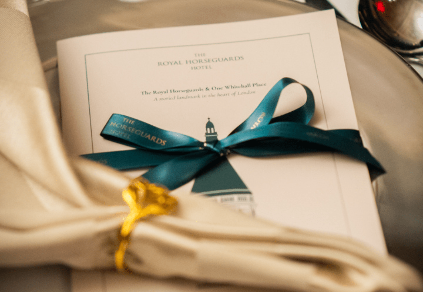 A folded cream-colored napkin with a gold napkin ring rests beside a booklet tied with a teal ribbon, featuring “The Royal Horseguards Hotel” printed on the cover.