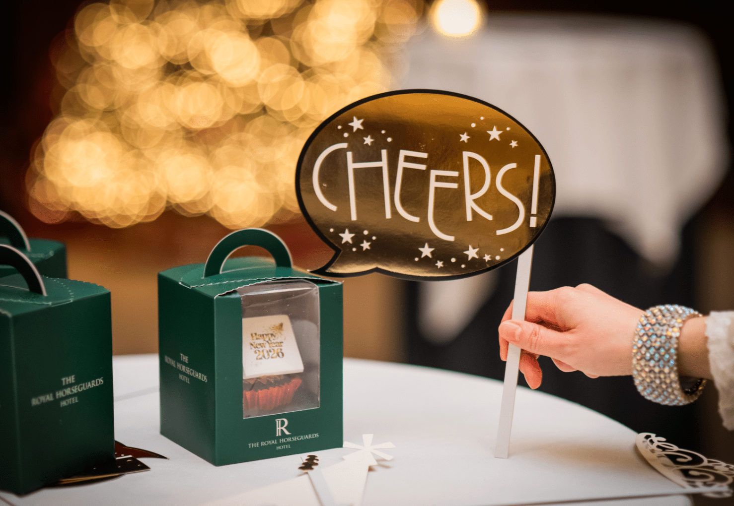 A hand holding a sign that says “CHEERS!” near a table with small green gift boxes, with warm blurred lights in the background.