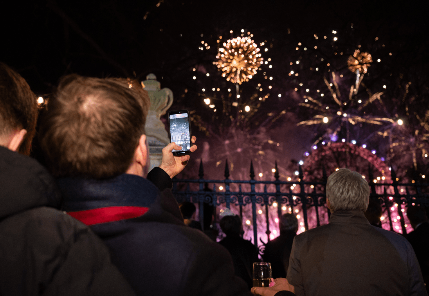 A crowd of people watching a nighttime fireworks display, with someone in the foreground holding up a phone to record the scene.